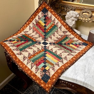 Könnte beinhalten: Ein quadratischer Stoffquilt mit einem Log-Cabin-Muster und einem dunkelblauen Quadrat in der Mitte. Das Design enthält Stoffstreifen in Rot, Gelb, Grün und Creme mit Blumen- und geometrischen Mustern, abgeschlossen mit einem breiten orangefarbenen Rand.