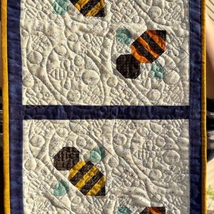 May include: A quilted wall hanging featuring a repeating pattern of bees. The bees are made of black, yellow, and orange fabric. The background is white with a floral pattern, and the edges are trimmed with blue and gold fabric.