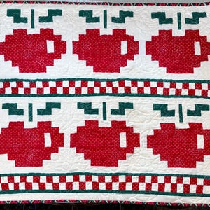 May include: A quilted square featuring a repeating pattern of red apple shapes with green stems on a white background. The apples have a dotted texture. Green and white checkered bands and a red and green border complete the design. The quilt is likely handmade.