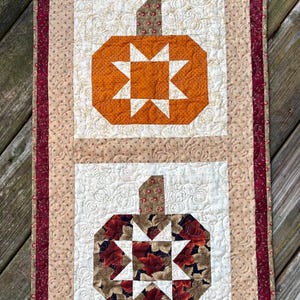 May include: A quilted wall hanging featuring two pumpkin designs. The top pumpkin is orange with a white starburst center, and the bottom pumpkin is a floral pattern with a white starburst center. The border is a mix of beige, burgundy, and white.