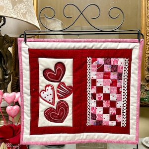 May include: A Valentine's Day themed quilted wall hanging. The piece features a red and white color scheme with appliqued hearts and a patchwork design. The edges are finished with a pink border.