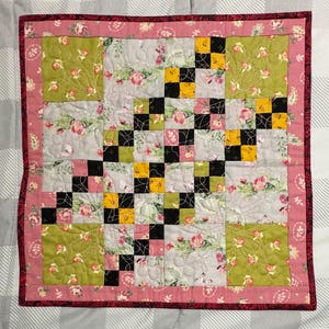 May include: A square patchwork quilt featuring a border of pink floral fabric. The center consists of alternating blocks of light gray floral fabric, solid olive green floral squares, and a grid of small black and yellow checkered squares.