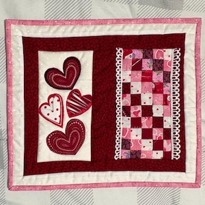May include: A rectangular fabric wall hanging featuring a deep red background with a white panel on the left displaying four appliqued hearts in red and white patterns. A vertical patchwork strip of pink and red squares with white lace trim.