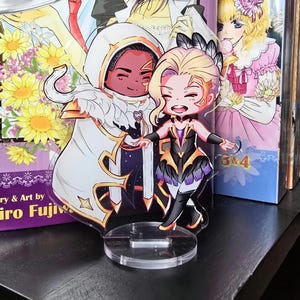 May include: Acrylic anime character standees featuring two figures with detailed outfits and expressions. The standees are placed in front of manga volumes with colorful illustrations. The background includes manga covers and a shelf.