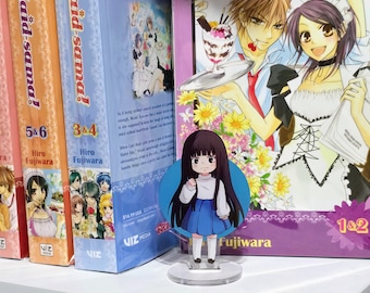 May include: A shelf displays several volumes of the manga series "Maid-sama!" with colorful covers. A clear acrylic figure of a character from the series stands in front. The figure is a girl in a white top and blue skirt.