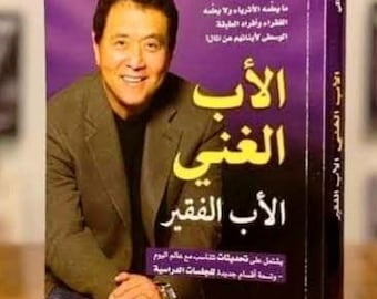 May include: A hardcover book titled "Rich Dad Poor Dad" in Arabic. The cover features a smiling man in a suit jacket against a purple background. The book's title is in large yellow letters, with additional text in Arabic.