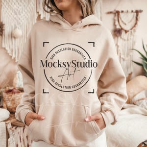 May include: A beige hoodie with a kangaroo pocket and a drawstring hood. The hoodie features a circular logo with the text "MocksyStudio Art" and "High Resolution Guaranteed". The model is wearing the hoodie.