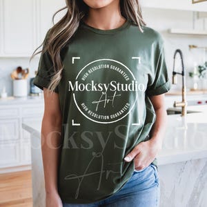 May include: An olive green t-shirt with rolled-up sleeves. The shirt features a white circular logo with the text "MocksyStudio Art" and "High Resolution Guaranteed". The model is wearing blue jeans.