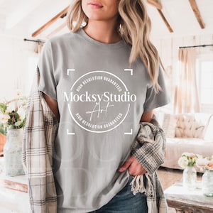 May include: A woman wearing a gray t-shirt with a white circle logo that says "MocksyStudio Art" and "High Resolution Guaranteed". She is wearing a plaid shirt over the t-shirt and blue jeans.