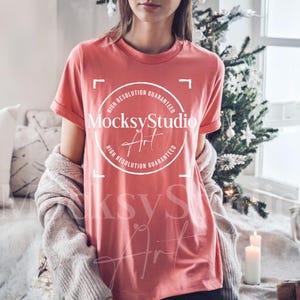 May include: A coral-colored t-shirt with rolled sleeves. The shirt features a white circular graphic with the text "MocksyStudio Art" and "High Resolution Guaranteed". A light-colored sweater is draped over the shoulders. A Christmas tree and candles are in the background.