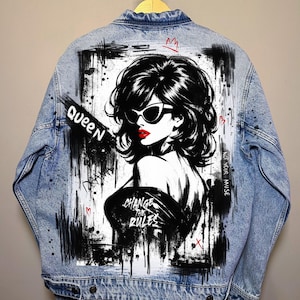 May include: Pale blue denim jacket with a black and white illustration of a woman in sunglasses and red lipstick. The jacket displays the words "QUEEN" and "CHANGE THE RULES" in black. Red heart and crown details are also present.