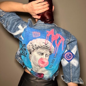 May include: A blue denim jacket with a painted design featuring a bust of a man blowing a bubble, a crown, and the text "bla bla bla".