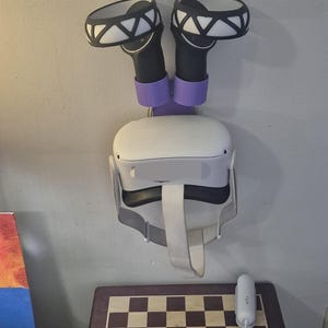May include: A white VR headset with a black strap and two black controllers with purple holders mounted on a white wall. The controllers have black and white geometric designs on the front. A white chessboard is on a table below the headset.