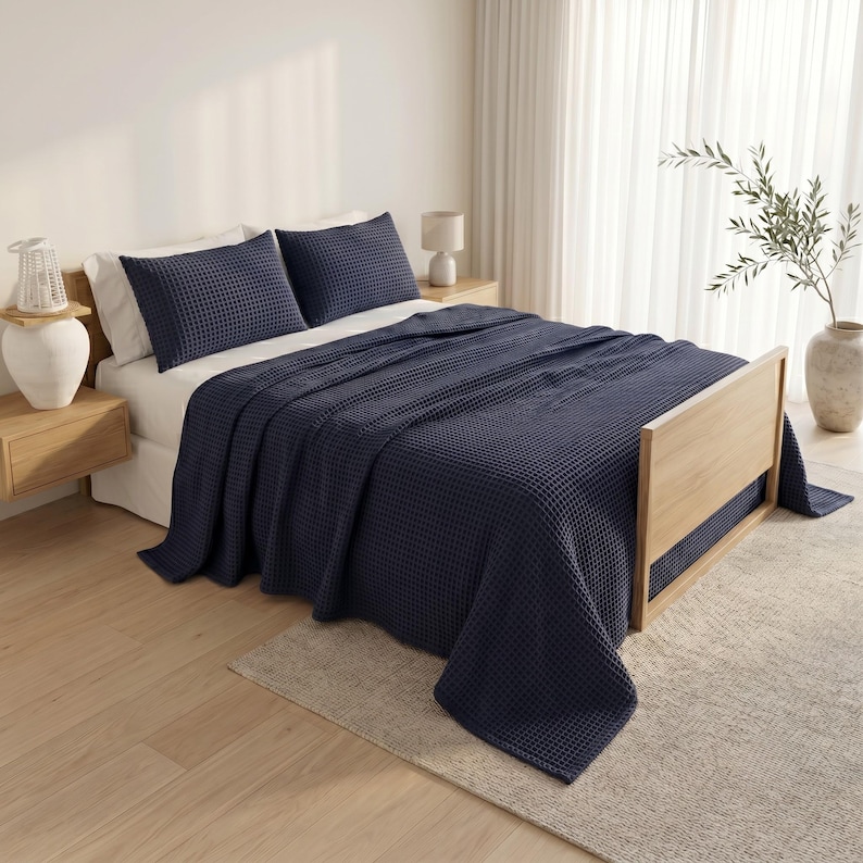 Op de afbeelding: Een donker marineblauwe sprei met wafelstructuur gedrapeerd over een houten bedframe. De set bevat twee bijpassende donker marineblauwe kussenslopen met wafelstructuur die op witte kussens rusten. Het beddengoed heeft een duidelijke rasterachtige textuur.