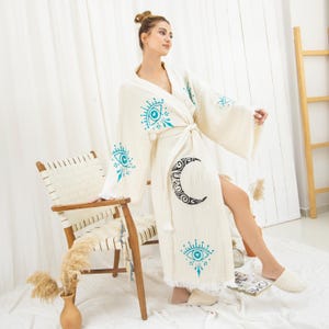 May include: Cream-colored robe with a wrap-around design, featuring turquoise eye and crescent moon patterns. The robe has wide sleeves and a fringed hem. A matching belt cinches the waist. The model is wearing the robe.