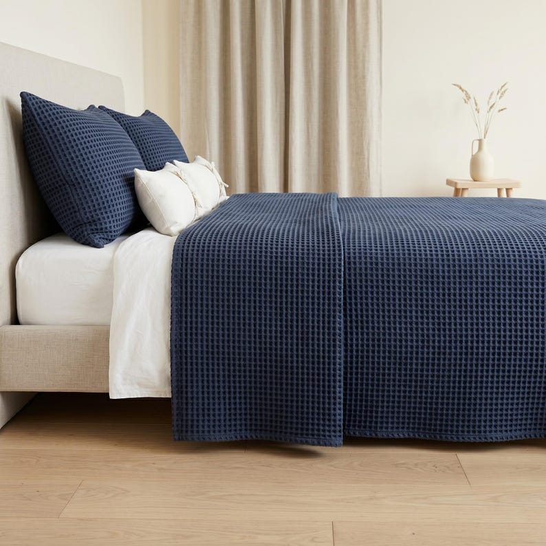 Op de afbeelding: Een donker marineblauwe bedsprei met wafelstructuur bedekt een bed met een wit laken en twee donker marineblauwe kussenslopen met wafelstructuur. Twee kleinere witte rechthoekige kussens liggen voor de donkerblauwe kussenslopen.