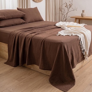 May include: A solid dark brown bed sheet set featuring a textured, crinkled fabric finish. The set includes two matching pillows and a large bed cover draped over a wooden bed frame, complemented by a cream-colored throw blanket with fringe edges.