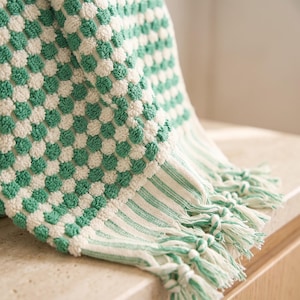 May include: A woven textile featuring a textured checkerboard pattern in alternating sage green and off-white squares. The fabric includes a striped border section at the bottom edge, finished with long, twisted fringe tassels in matching green and white.