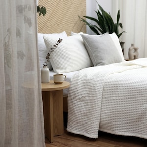 Op de afbeelding: Een witte bedsprei met wafelstructuur en bijpassende kussenslopen liggen op een bed. Een lichtgrijs vierkant kussen ligt op het bed. Een ronde houten bijzettafel bevat een kleine keramische vaas met een takje en een kleine keramische mok.
