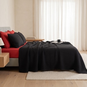 Op de afbeelding: Een bed met een zwarte wafelstructuur deken, twee zwarte wafelstructuur kussenslopen en twee effen rode kussenslopen. Het bed is opgemaakt met een rood hoeslaken en een kleine zwarte mok staat op de deken bij het hoofdeinde van het bed.