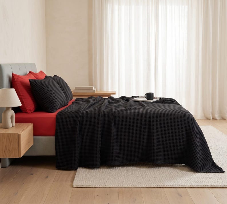 Op de afbeelding: Een bed met een zwarte wafelstructuur deken, twee zwarte wafelstructuur kussenslopen en twee effen rode kussenslopen. Het bed is opgemaakt met een rood hoeslaken en een kleine zwarte mok staat op de deken bij het hoofdeinde van het bed.
