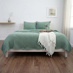 Op de afbeelding: Een saliegroene sprei met wafelstructuur bedekt een houten bedframe, vergezeld van twee bijpassende rechthoekige kussens. Een cr&egrave;mekleurige plaid met decoratieve kwastjes is over het voeteneinde van het bed gedrapeerd voor extra textuur.