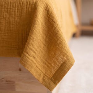 May include: A mustard yellow bedspread or throw blanket made of a crinkled, multi-layered fabric. The textile features a visible grid-like texture and a stitched hem border along the edges, showcasing a soft, lightweight, and semi-sheer appearance.
