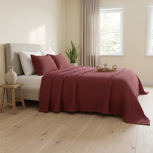 May include: A deep red waffle-weave bedspread covers a mattress on a light gray upholstered bed frame. Two matching red waffle-weave pillow shams rest against white pillows. A small woven tray sits on the bed, and a ceramic vase stands on a wooden stool.