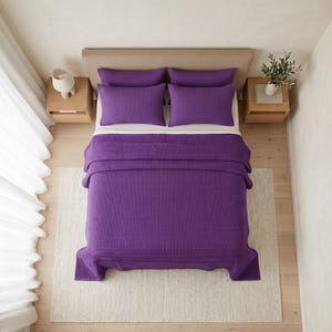 Op de afbeelding: Een bed met een dieppaarse deken met wafelstructuur en twee bijpassende rechthoekige kussens. Het beddengoed ligt op een matras in neutrale tinten met een beige hoofdeinde, geflankeerd door twee nachtkastjes van licht hout.