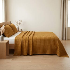 Op de afbeelding: Een mosterdgele sprei met wafelstructuur bedekt een bed met twee bijpassende vierkante kussens. Het beddengoed staat tegen een effen muur met een lichte houten vloer zichtbaar onder het bedframe en een houten nachtkastje naast het bed.