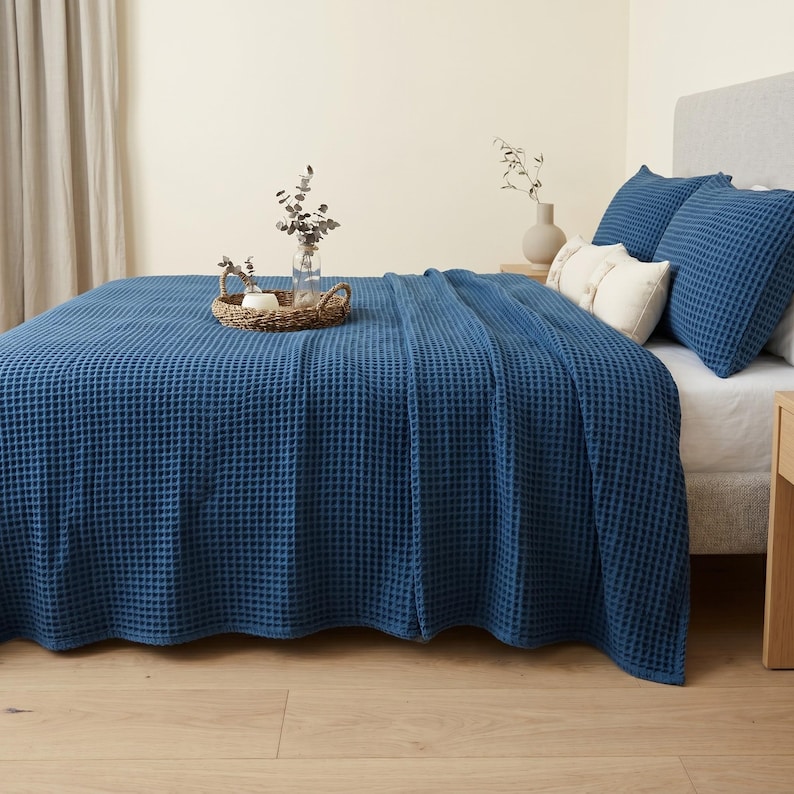 Op de afbeelding: Een diepblauwe sprei met wafelstructuur bedekt een bed, vergezeld door twee bijpassende blauwe kussenslopen en twee kleinere cr&egrave;mekleurige sierkussens. Een gevlochten mand met een glazen vaas met gedroogde eucalyptusstengels staat op de sprei.