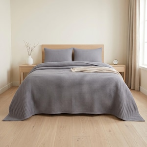 Op de afbeelding: Een grijze bedsprei met wafelstructuur op een bed met twee bijpassende rechthoekige kussens. Een gevouwen cr&egrave;mekleurige plaid ligt bovenop de sprei. Het bed staat tegen een effen muur met een houten hoofdeinde en frame.