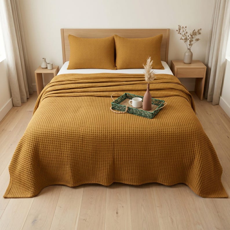 Op de afbeelding: Een mosterdgele sprei met wafelstructuur op een bed met twee bijpassende kussens. Op het bed staat een groen rechthoekig dienblad met een witte keramische mok en een bruine vaas met een gedroogde bloemstengel, wat een textuurrijk geheel vormt.
