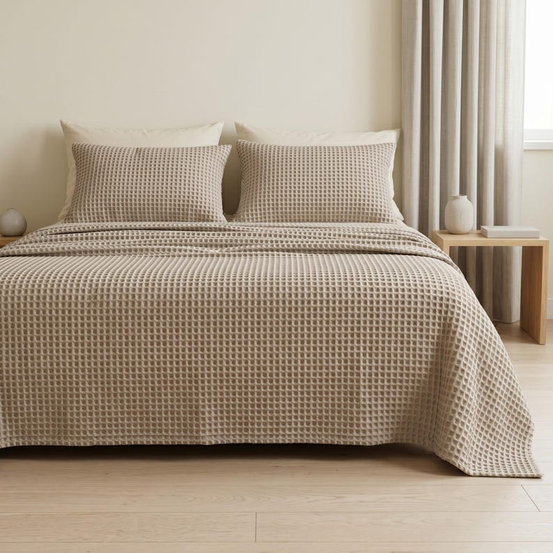 Op de afbeelding: Een beige bedsprei met wafelstructuur die een bed bedekt met twee bijpassende rechthoekige kussens. De stof heeft een duidelijke rasterachtige textuur over het gehele oppervlak van het beddengoed, wat zorgt voor een uniforme uitstraling.
