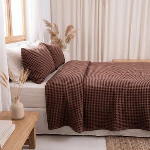 Op de afbeelding: Een donkerbruine wafelstructuur sprei en twee bijpassende kussenslopen op een bed met wit beddengoed. Het beddengoed heeft overal een duidelijke rasterachtige textuur, wat zorgt voor een uniforme uitstraling op de kussens en de deken.