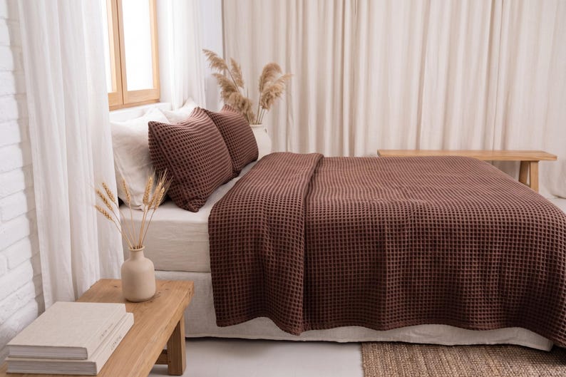 Op de afbeelding: Een donkerbruine wafelstructuur sprei en twee bijpassende kussenslopen op een bed met wit beddengoed. Het beddengoed heeft overal een duidelijke rasterachtige textuur, wat zorgt voor een uniforme uitstraling op de kussens en de deken.