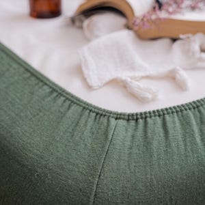 May include: A deep olive green fitted sheet featuring a textured, woven fabric surface with a gathered elastic edge. The fabric displays a visible, fine-grained weave pattern, providing a matte finish to the material across the corner seam.