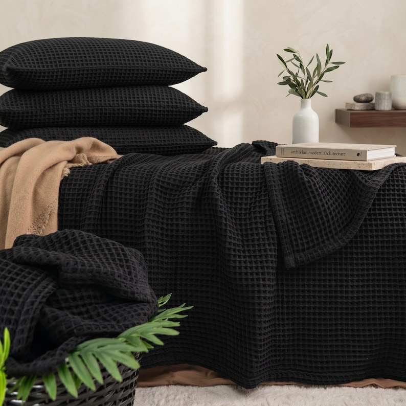Op de afbeelding: Een set zwart beddengoed met wafelstructuur, bestaande uit drie gestapelde kussens en een grote deken die over een oppervlak is gedrapeerd. Een beige plaid ligt onder het zwarte beddengoed en een wit boek ligt bovenop de deken.