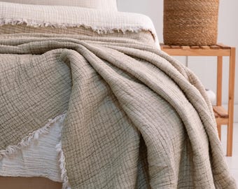 May include: A soft, beige throw blanket with a textured weave and frayed edges. The blanket is folded and draped over a bed.