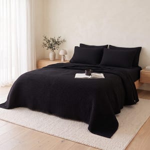 Op de afbeelding: Een effen zwarte bedsprei met wafelstructuur die een matras bedekt, vergezeld van twee bijpassende rechthoekige kussenslopen. De gestructureerde stof heeft een rasterachtig patroon, wat zorgt voor een uniforme uitstraling van de beddengoedset.