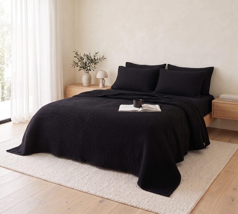 Op de afbeelding: Een effen zwarte bedsprei met wafelstructuur die een matras bedekt, vergezeld van twee bijpassende rechthoekige kussenslopen. De gestructureerde stof heeft een rasterachtig patroon, wat zorgt voor een uniforme uitstraling van de beddengoedset.