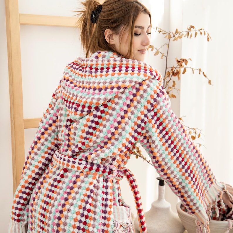 May include: A colorful hooded robe with a geometric pattern in shades of pink, red, orange, green, and white. The robe has a tie belt and fringe trim.