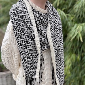 Puede incluir: Bufanda con estampado en blanco y negro con flecos, sobre un suéter de punto trenzado color crema. La bufanda presenta un diseño geométrico y está hecha de un material suave. Un accesorio elegante para el invierno.