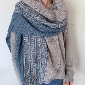 May include: A large, rectangular knitted shawl featuring a two-tone colour scheme of dusty blue and beige. The fabric displays a textured, moss-stitch pattern combined with a geometric, interlocking maze-like design that runs along the edges.