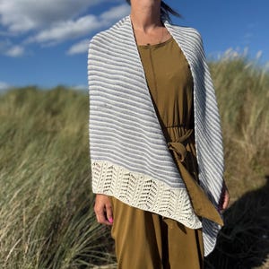 May include: A white and grey striped shawl with a cream-coloured, openwork border. The shawl is draped over a person wearing a mustard-coloured dress, with a background of tall grass and a blue sky.