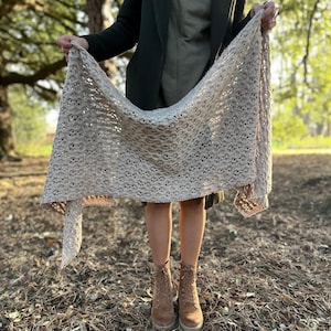 Puede incluir: Un chal de ganchillo beige con un delicado patrón de calado. El chal está drapeado sobre los hombros de una persona y sostenido en sus manos.