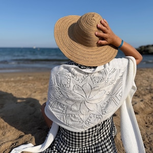 Könnte beinhalten: Eine Person trägt einen Sonnenhut aus Stroh und einen weißen Schal mit floralem Spitzendesign, der an einem Sandstrand sitzt. Der Schal ist über einem schwarz-weiß karierten Kleid drapiert. Ein blaues Armband ist sichtbar.
