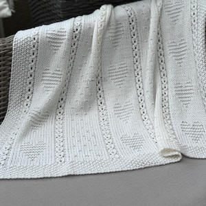 May include: A white knitted blanket with a heart pattern. The blanket features a detailed design with rows of hearts and decorative borders. The soft, textured blanket is draped over a wicker chair, suggesting a cozy and comforting feel.