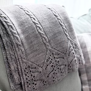 May include: A folded gray knitted blanket with cable knit and lace detailing. The blanket is folded over a gray surface, showcasing the intricate patterns and textures of the knitwear. The blanket's edges feature a delicate, openwork lace design.