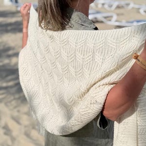 Puede incluir: Un chal de punto color blanco roto con un patrón repetitivo de trenzas verticales en forma de hoja. El tejido tiene una apariencia suave y texturizada con una estructura de punto consistente, mostrando un diseño detallado y en relieve.